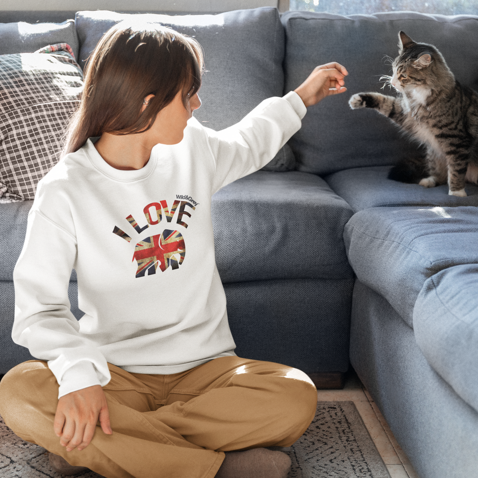 Child wearing a sweatshirt with 'I love' and a Union Jack design, sitting on the floor next to a cat on a couch.