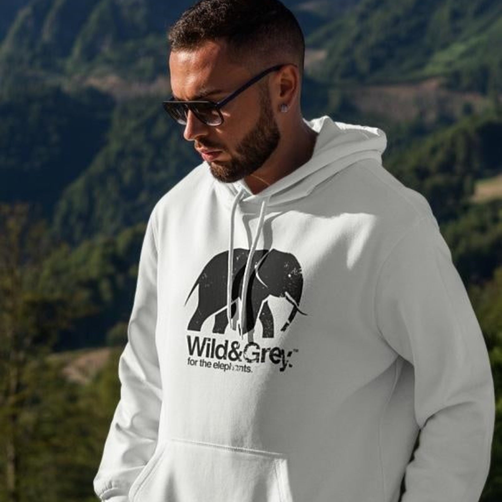 A man wearing sunglasses and a white hoodie with an elephant tusker design and the brand 'Wild&Grey' on it, standing outdoors with a mountainous landscape in the background.